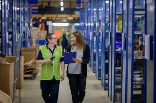 Two warehouse staff walking between storage racks while reviewing a tablet